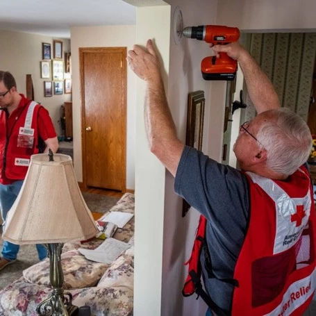 A man in a Red Cross vest with gray t-shirt holding a drill to a door frame, another man in a Red Cross shirt and jeans walking on the other side of the door.
